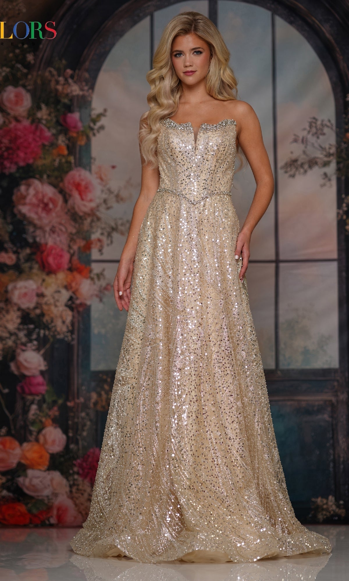 A woman stands before flowers and arched windows, wearing the Colors Dress 3790—a long strapless prom dress adorned with gold sequins that shimmer with elegant style.